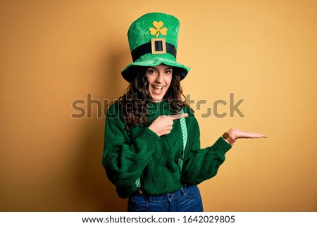Beautiful curly hair woman wearing green hat with clover celebrating saint patricks day amazed and smiling to the camera while presenting with hand and pointing with finger.