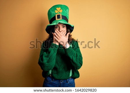 Beautiful curly hair woman wearing green hat with clover celebrating saint patricks day shocked covering mouth with hands for mistake. Secret concept.