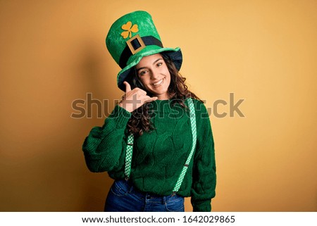 Beautiful curly hair woman wearing green hat with clover celebrating saint patricks day smiling doing phone gesture with hand and fingers like talking on the telephone. Communicating concepts.