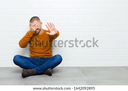 young handsome man covering face with hand and putting other hand up front to stop camera, refusing photos or pictures sitting on the floor