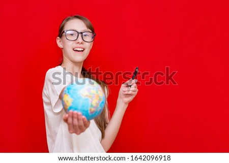 Amazed caucasian young girl with glasses show a globe.Education concept, back to school, education, young teenage schoolgirl.Red studio background,Copy space.