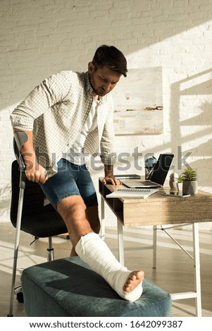 Man with broken leg standing near table with laptop and notebooks