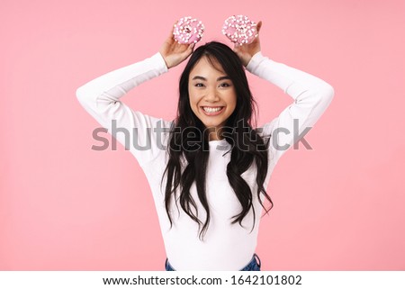 Image of young brunette asian woman with long hair smiling and holding donuts isolated over pink background