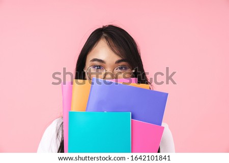 Image of young brunette asian student girl wearing eyeglasses holding paper folders isolated over pink background
