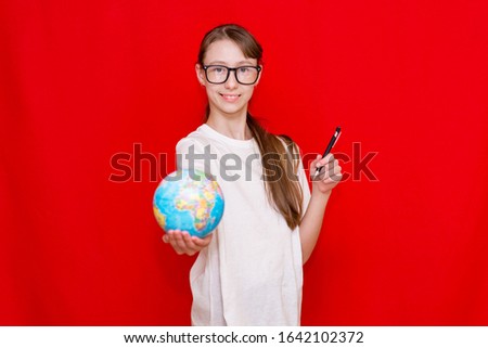 Young smiling schoolgirl in glasses thinking something.Shows to globe with pen.Bright red studio background. Copy space.