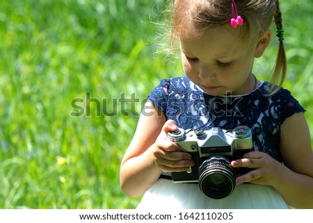 A little girl takes a picture with a retro film camera in the summer in the park. Little pretty child baby girl in light dress holding retro vintage photo camera on green grass in city park.