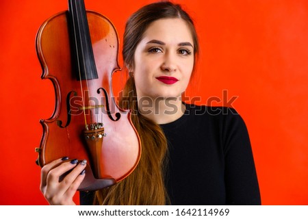 Portrait of a pretty brunette musician girl with a smile in a black dress on a red background holds a violin in her hands