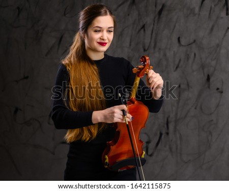 Portrait of a pretty brunette musician girl in a black dress on a gray background holds a violin in her hands