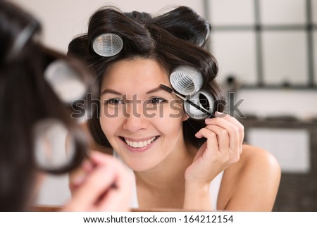 Woman in hair curlers applying eye makeup in the bathroom mirror contouring her eyebrows with eyeliner