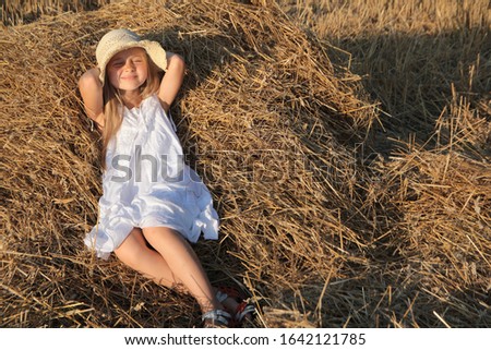 Little girl in a white dress plays in the meadow. Girl throws hay into the air.Little girl in a white hat.