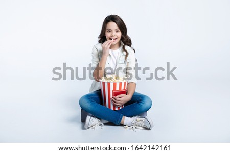 Favorite treat. Petite girl with a big tube of tasty popcorn is sitting in a lotus position, looking in the camera and eating.