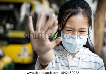 Asian child girl wearing protective mask at bus stop in the  city,show stop gesture,hand"NO"sign,people against air pollution,smog,dust allergy,PM2.5,air contamination,polluted environment from a car