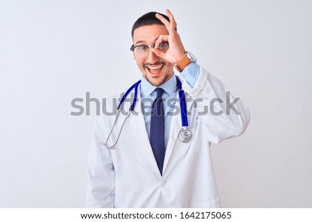 Young doctor man wearing stethoscope over isolated background doing ok gesture with hand smiling, eye looking through fingers with happy face.