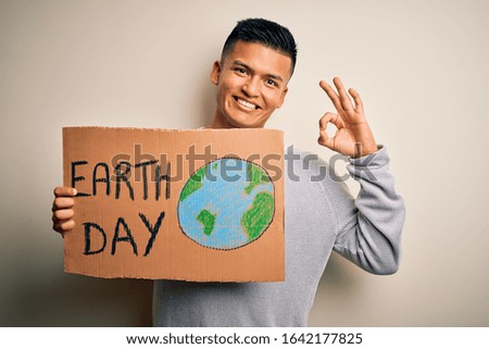 Young handsome latin man holding banner asking for earth and enviroment conservation doing ok sign with fingers, excellent symbol