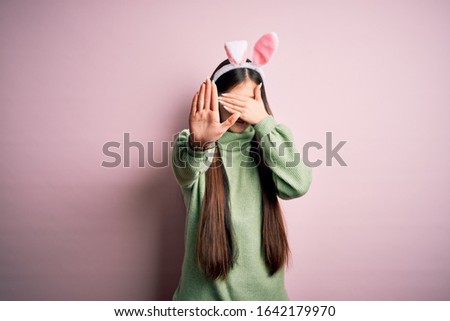 Young asian woman wearing cute easter bunny ears over pink background covering eyes with hands and doing stop gesture with sad and fear expression. Embarrassed and negative concept.