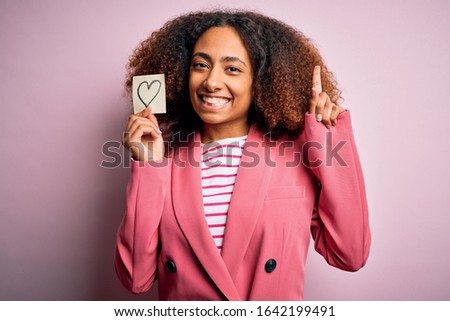 Young african american woman with afro hair holding paper with heart over pink background surprised with an idea or question pointing finger with happy face, number one