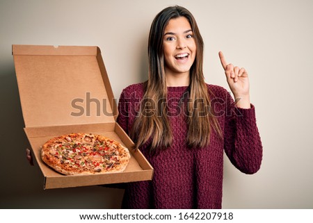 Young beautiful girl holding delivery box with Italian pizza standing over white background surprised with an idea or question pointing finger with happy face, number one