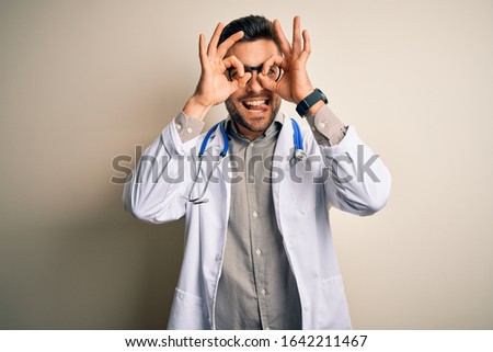 Young doctor man wearing glasses, medical white robe and stethoscope over isolated background doing ok gesture like binoculars sticking tongue out, eyes looking through fingers. Crazy expression.