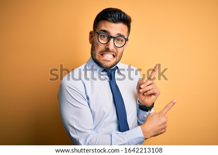 Young handsome businessman wearing tie and glasses standing over yellow background Pointing aside worried and nervous with both hands, concerned and surprised expression