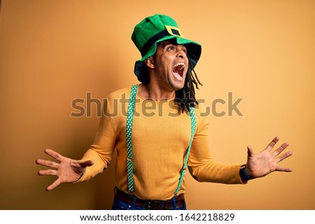 Young african american man wearing green hat celebrating saint patricks day crazy and mad shouting and yelling with aggressive expression and arms raised. Frustration concept.
