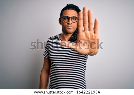 Young handsome african american man with dreadlocks wearing striped t-shirt and glasses doing stop sing with palm of the hand. Warning expression with negative and serious gesture on the face.