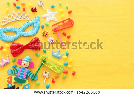 Party colorful noisemaker, mask and cute clown doll over yellow wooden background . Top view, flat lay