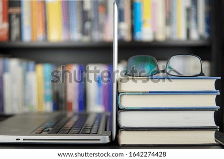 Laptop computer and a stack of books on a working table and a bookshelf in background