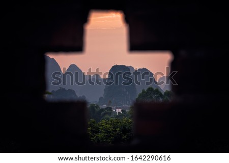 Beautiful impressive karst mountain landscape in Yangshuo at dusk seen through the cross shaped hole in a brick boundary wall, Guangxi Province, China