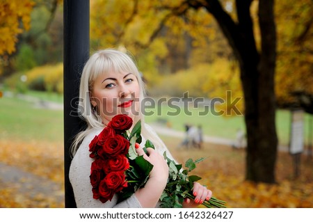Portrait of a sweet, plump, middle-aged woman who looks at the camera with irony, standing in nature against a background of yellow trees with a bouquet of red roses in her hands. Closeup.