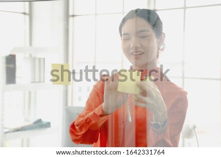 Brainstorming Concept. Asia businesswoman writing memo on sticky notes on glass wall at office, free space
