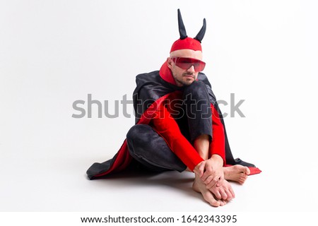 Fashionable man in black red diabolical carnival clothes wearing devil horns and sportive sunglasses and posing with confidence over white studio background.