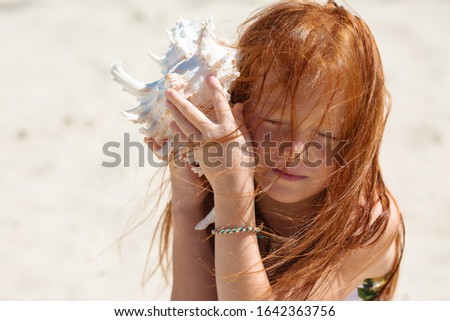 little girl with red hair sits on a sandy beach and holds a large seashell near her ear, space for text Royalty-Free Stock Photo #1642363756