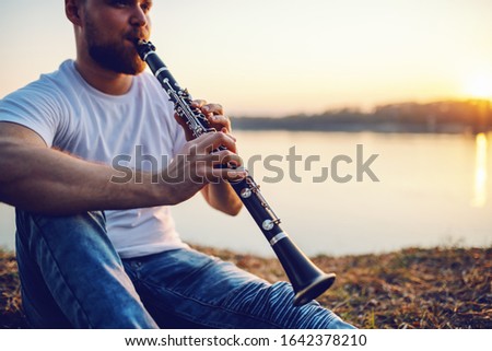 Young handsome bearded caucasian blond man sitting on cliff and playing clarinet. In background is river and sunset.