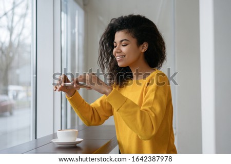African American woman food blogger taking picture of coffee on her smartphone, sitting in modern cafe. Portrait of happy emotional social media influencer using mobile phone, streaming video online