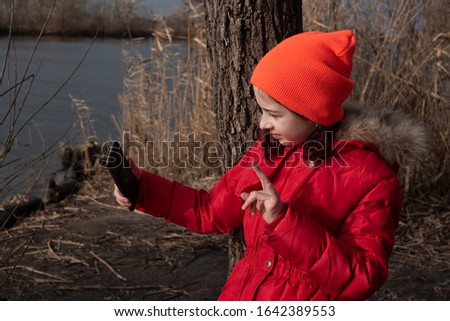 Girl dressed in red jacket, poses for making selfie or photo of herself with smartphone. People and leisure concept. Girl makes selfie in a red jacket.Girl 9 years old with a smartphone in her hands.