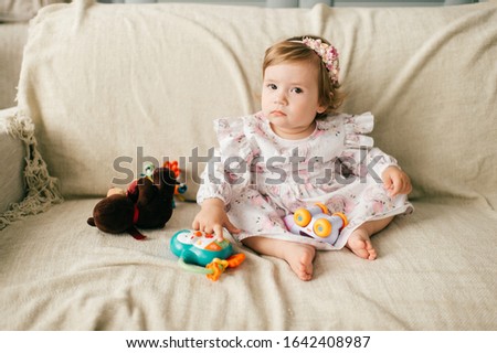 Little cute caucasian girl in attractive dress sits on the couch, plays with her various toys and smiles