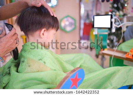 Cute worried little Asian 3 - 4 years old toddler boy child getting a haircut while watching cartoon on smartphone in modern hairdresser's barber shop at department store in Bangkok Thailand