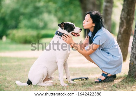 Young caucasian female with black hair in blue dress sits with her lovely black-and-white dog in the park and smiles