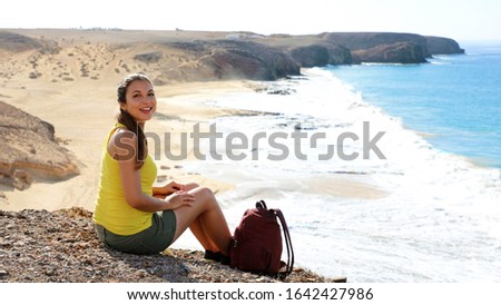 Beautiful young backpacker sitting enjoying view of the ocean waves and looking at camera on Playa Papagayo beach in Lanzarote, Canary Islands, Spain