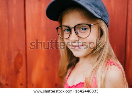 Picture of cute caucasian girl with long fair hair and in pink dress, glasses and denim cap smiles and makes faces against the wooden wall in village