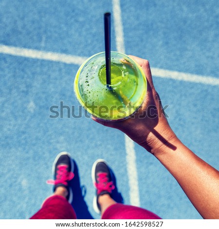 Green smoothie woman drinking plastic cup breakfast meal takeaway to go after morning run on blue tracks. Healthy lifestyle sporty person pov of hand holding glass with running shoes feet selfie.