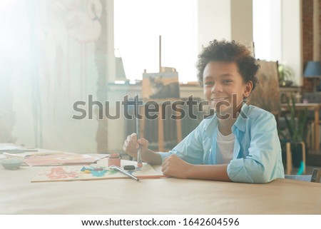 Portrait of African young boy smiling at camera while sitting at the table and drawing a picture with paints and brush