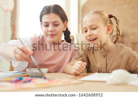 Two happy friends sitting at the table and painting a picture together with paints at school