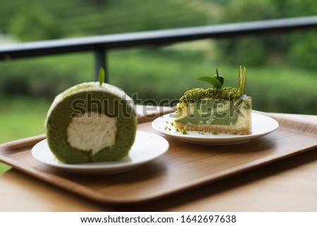 Slice of green tea cheesecake topped with green tea leaf on white plate with blur of green tea yam roll cake at Choui Fong Tea Plantation, Chiang Rai, Thailand. Selective focus image. Royalty-Free Stock Photo #1642697638