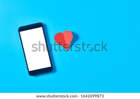 Black smartphone with isolated white screen for text, picture, photo and other graphics near red paper heart lies on blue table. Valentines day and love concept. Space for text. Top view