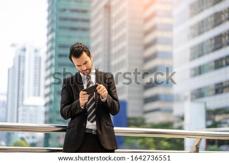 handsome caucasian businessman using and playing on smartphone smiling joyfully, representing relaxing and having fun, standing on a bridge platform with tall building and busy road in the background