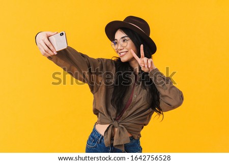 Image of young brunette asian woman wearing hat and eyeglasses taking selfie photo on cellphone isolated over yellow background