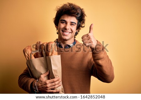 Young handsome man holding paper bag with bread standing over isolated yellow background happy with big smile doing ok sign, thumb up with fingers, excellent sign