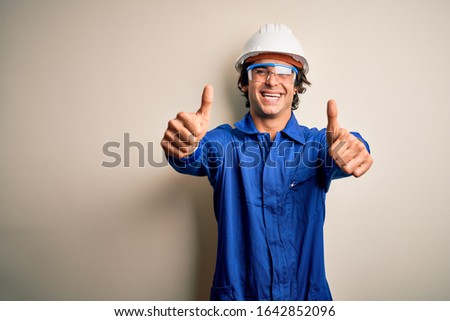 Young constructor man wearing uniform and security helmet over isolated white background approving doing positive gesture with hand, thumbs up smiling and happy for success. Winner gesture.