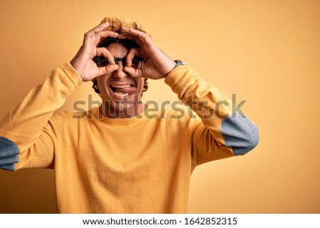 Young handsome man wearing casual t-shirt and glasses over isolated yellow background doing ok gesture like binoculars sticking tongue out, eyes looking through fingers. Crazy expression.